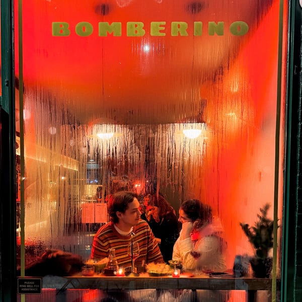 Steamy window view of two people dining inside Bomberino cafe, illuminated by warm red light.
