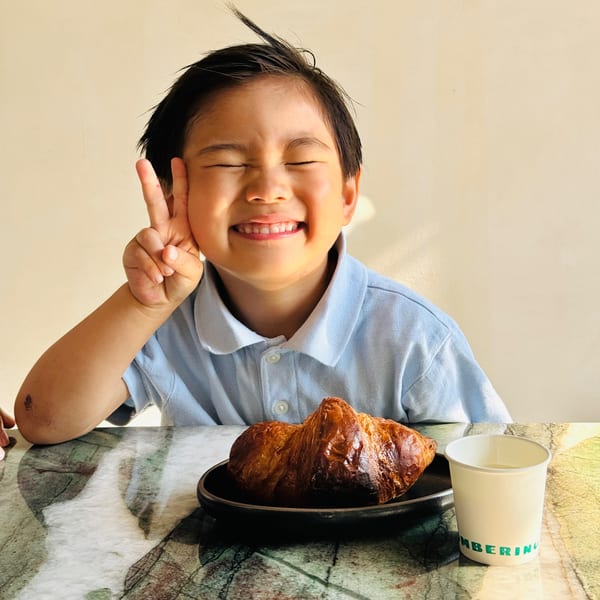 A happy child making a peace sign, with a croissant and Bomberino cup on a marble table.