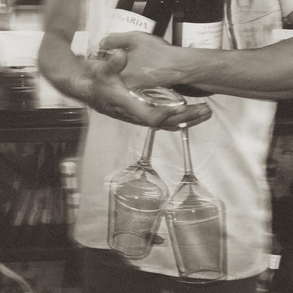 A server's hands holding two wine glasses upside down, with a bottle of wine in the background.