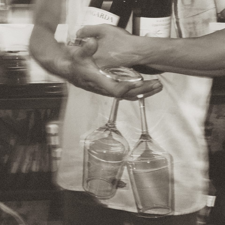 A server's hands holding two wine glasses upside down, with a bottle of wine in the background.