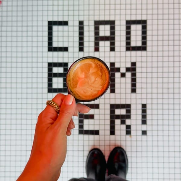 A hand holding a coffee cup over a tiled floor spelling "CIAO BOMBER!" with black boots visible.