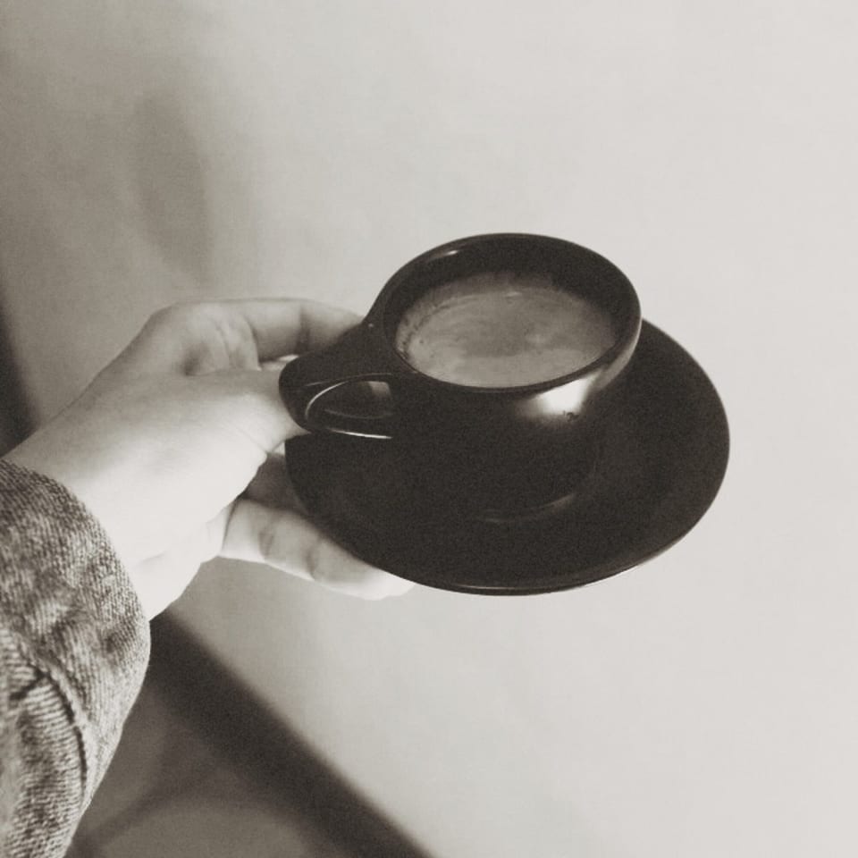 A hand holds a small black cup of espresso with a matching saucer in black and white.
