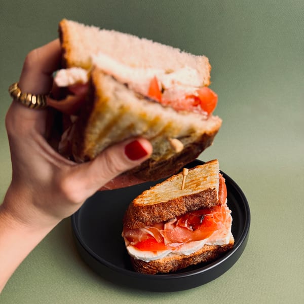 A hand holds a Bomberino cafe sandwich with prosciutto, mozzarella, and tomato over another on a black plate.