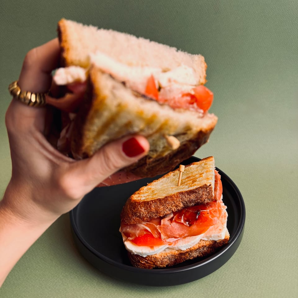 A hand holds a Bomberino cafe sandwich with prosciutto, mozzarella, and tomato over another on a black plate.