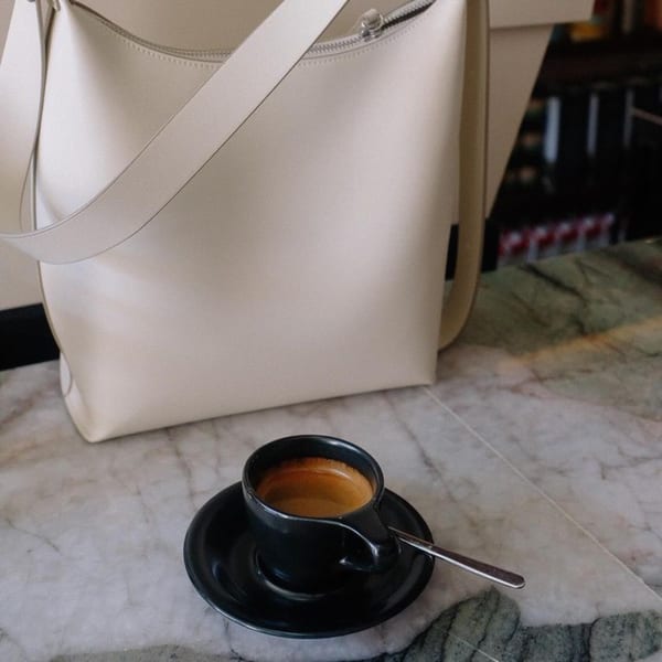 A black espresso cup with a spoon sits on a marble table next to a cream-colored bag.