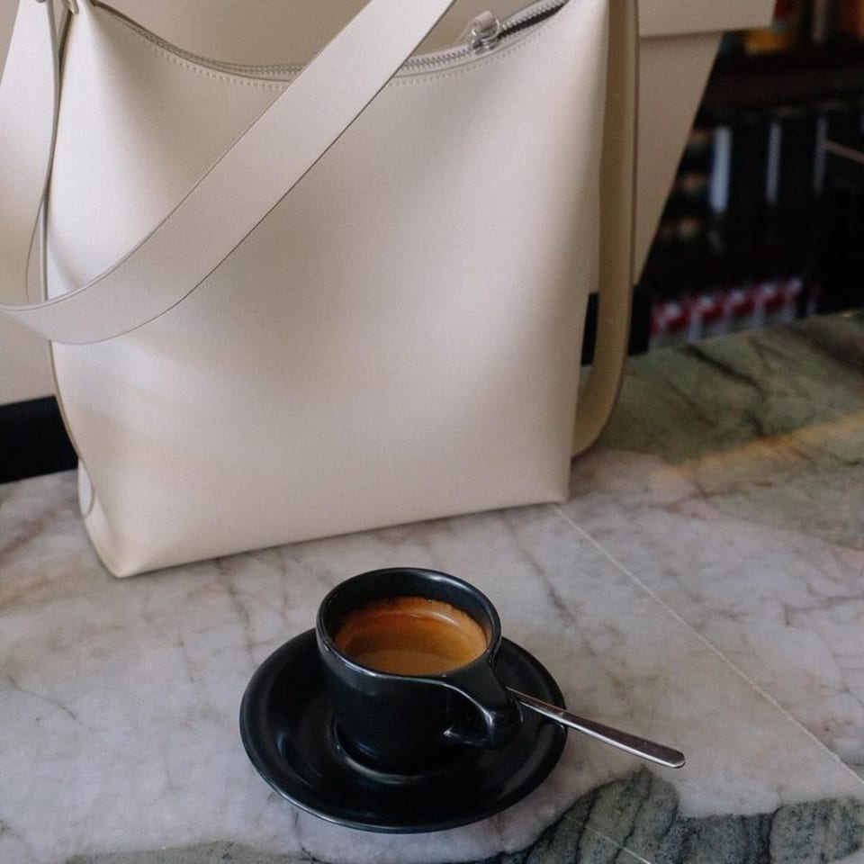 A black espresso cup with a spoon sits on a marble table next to a cream-colored bag.