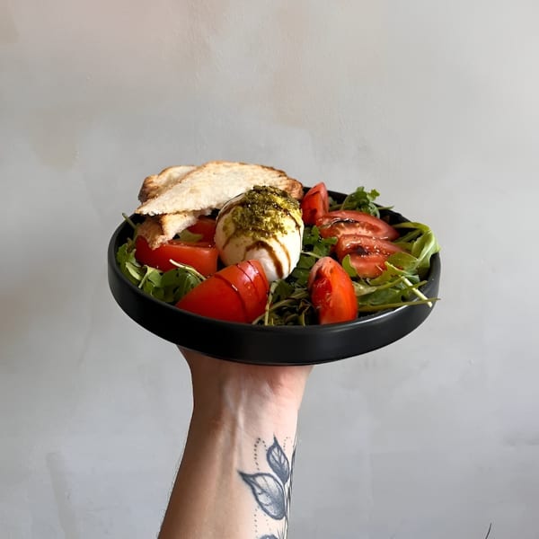 A hand holding a black plate with burrata, tomatoes, arugula, pesto, and toasted bread.