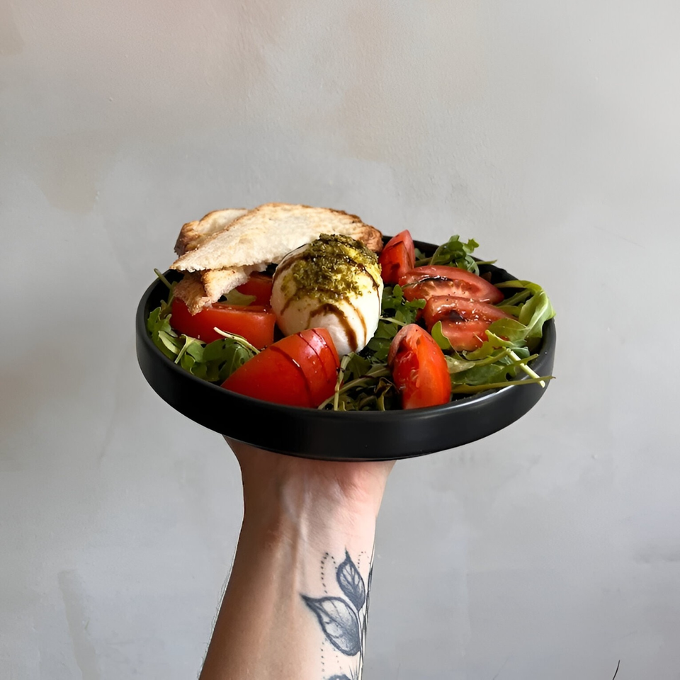A hand holding a black plate with burrata, tomatoes, arugula, pesto, and toasted bread.