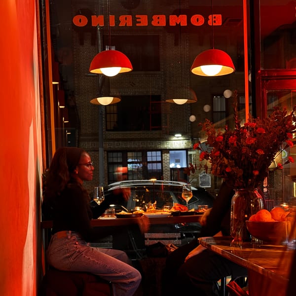 A woman in glasses sits at a table inside Bomberino cafe, illuminated by red lighting.