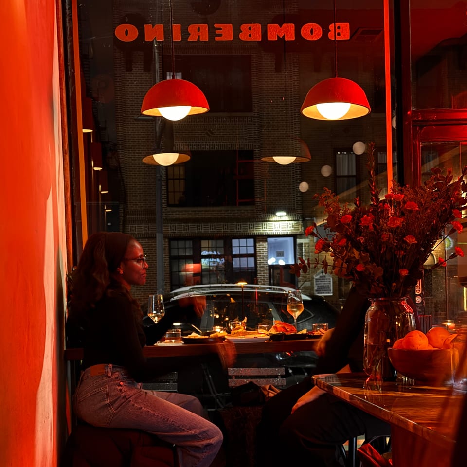 A woman in glasses sits at a table inside Bomberino cafe, illuminated by red lighting.