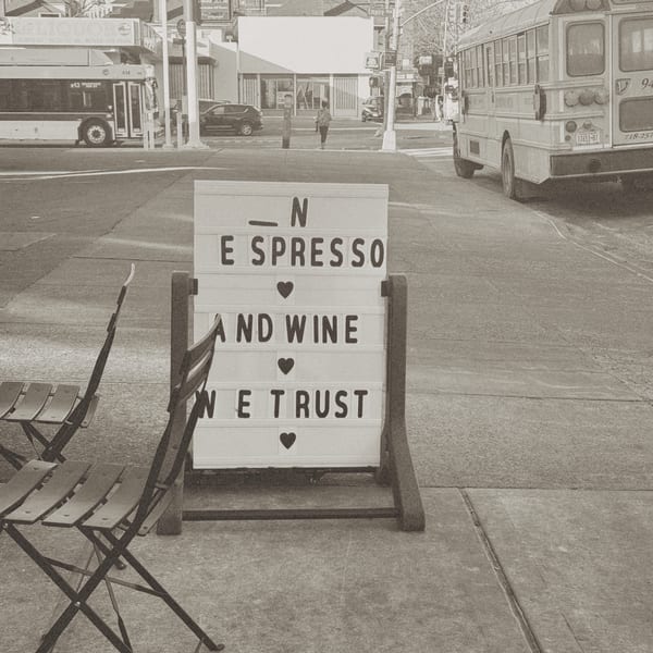 A Bomberino cafe sign reads "In Espresso and Wine We Trust" on a city sidewalk.