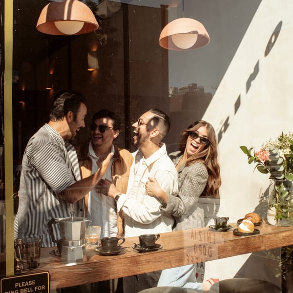 Four friends laughing inside Bomberino cafe, seen through a sunlit window with coffee and pastries.