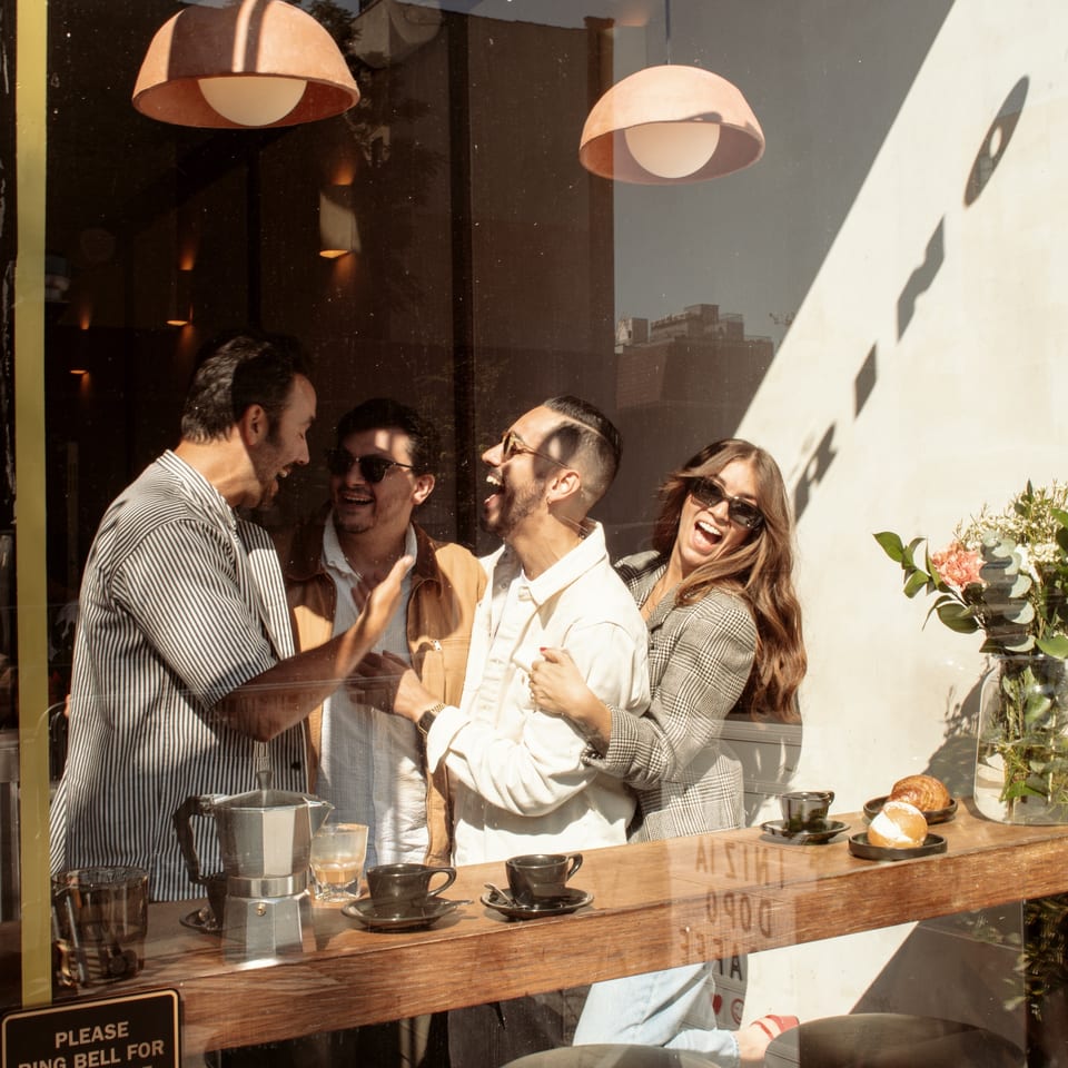 Four friends laughing inside Bomberino cafe, seen through a sunlit window with coffee and pastries.