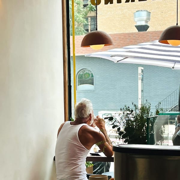 A man in a white tank top sips a drink at a cafe window, looking out at the street.