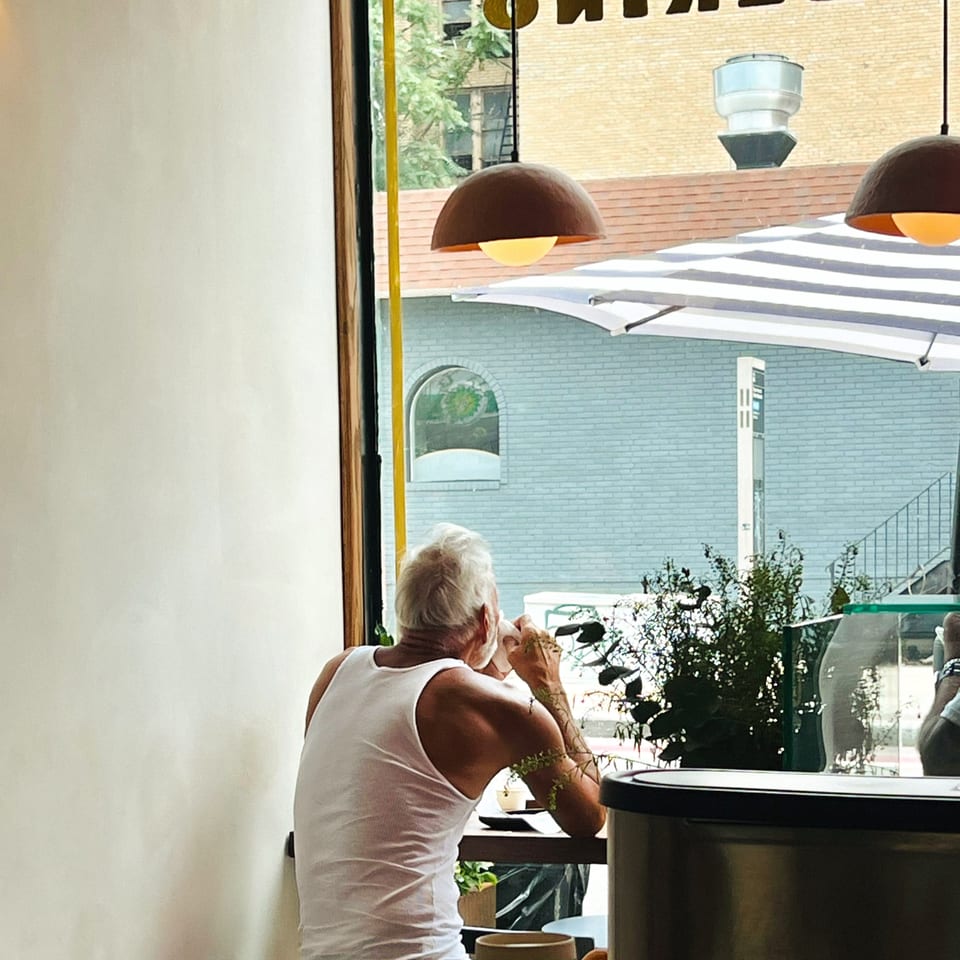 A man in a white tank top sips a drink at a cafe window, looking out at the street.