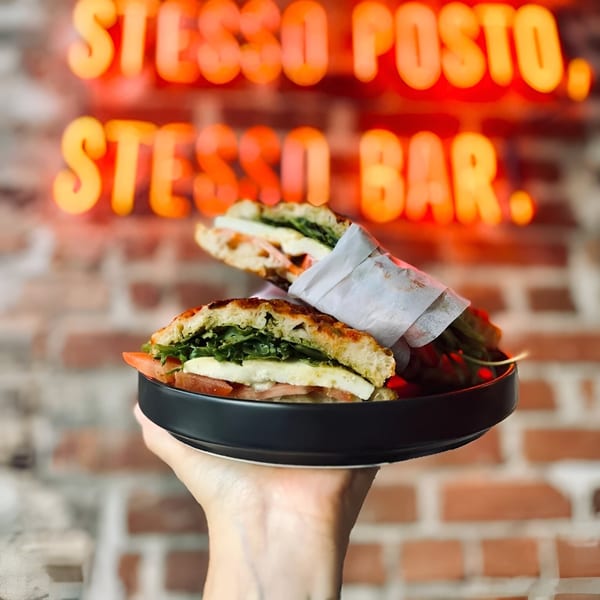 A hand holds a plate with two Bomberino cafe sandwiches against a brick wall with neon signs.